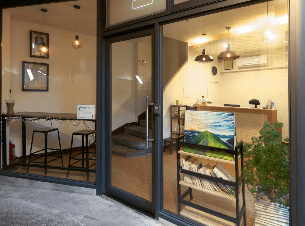 【瑞芳住宿】清芳民宿大廳與接待區，玻璃門外可見閱讀書架與畫作展示，內部設有櫃台與高腳桌座位區，燈光溫暖營造舒適迎賓氛圍。