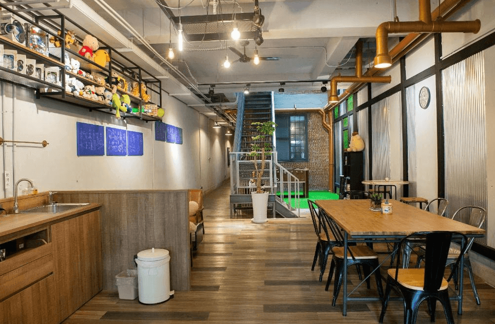 【台南寵物友善住宿】玩聚安平民宿的公共交誼廳，融合工業風與木質設計，設有長桌、廚房區與玩偶收藏展示櫃，空間溫馨且具設計感，適合旅人交流。