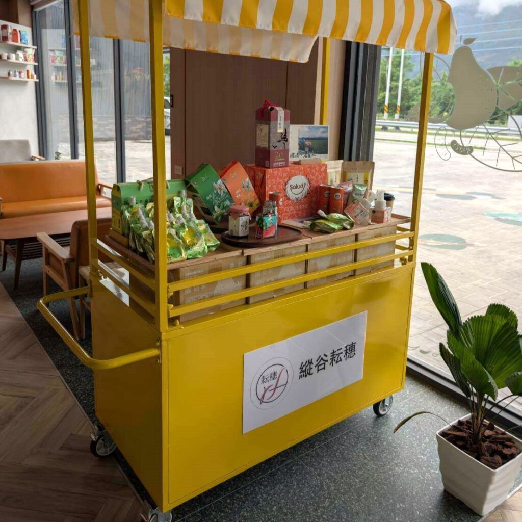【瑞穗美食】「縱谷耘穗咖啡廳」室內展示區，一台黃色推車上陳列多款在地農產品與伴手禮，包括果乾、茶包、飲品與蜂蜜等，搭配黃白條紋遮棚設計，整體氛圍明亮溫馨。