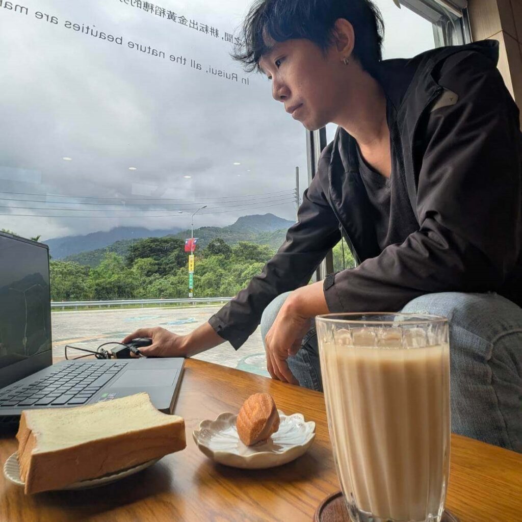 【瑞穗美食】在「縱谷耘穗咖啡廳」生活口袋創作者在窗邊用電腦，一旁桌上擺有鮮奶茶、厚片吐司與小點心，窗外可見遠方山景與雲霧，氛圍寧靜悠閒。