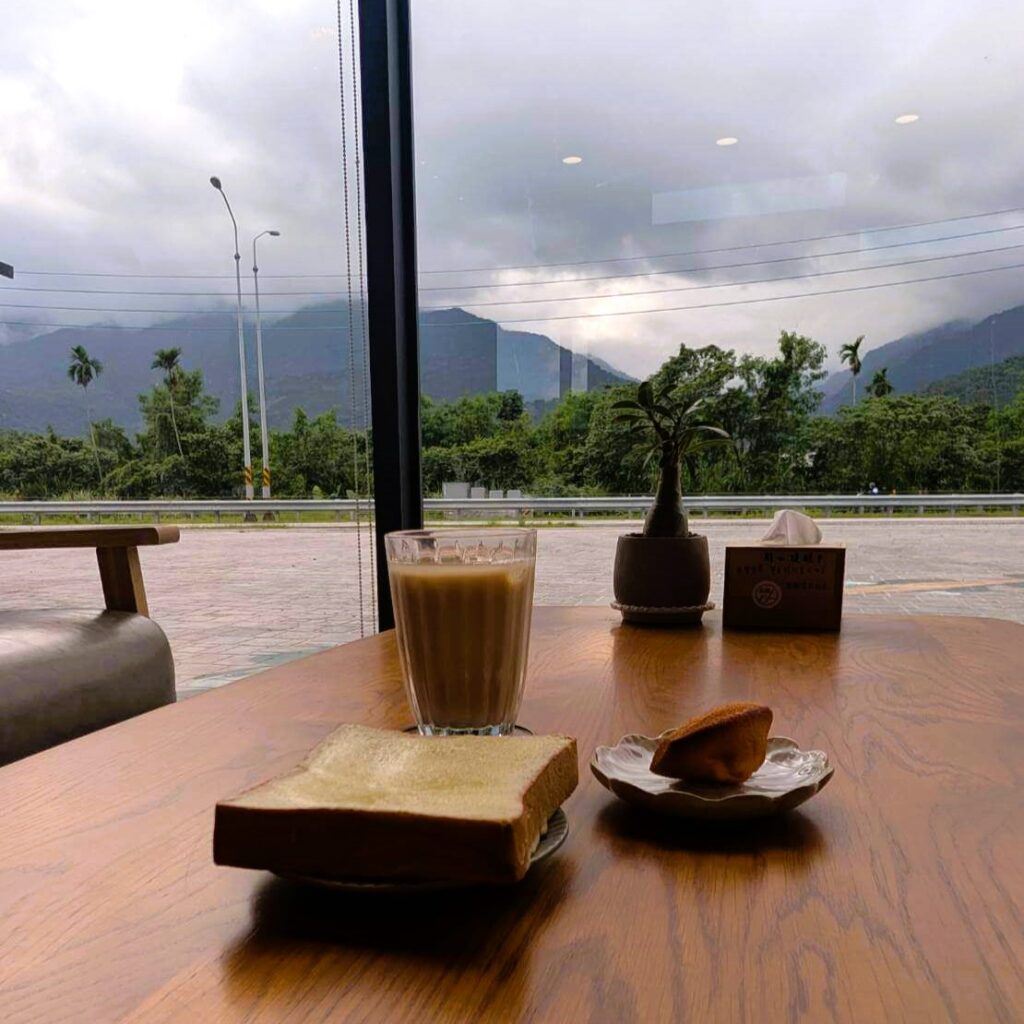 【瑞穗美食】「縱谷耘穗咖啡廳」窗邊桌面擺著一杯鮮奶茶、厚片吐司與小點心，背景為花蓮縱谷山景，透過大片玻璃窗能欣賞自然景色，氣氛愜意溫暖。