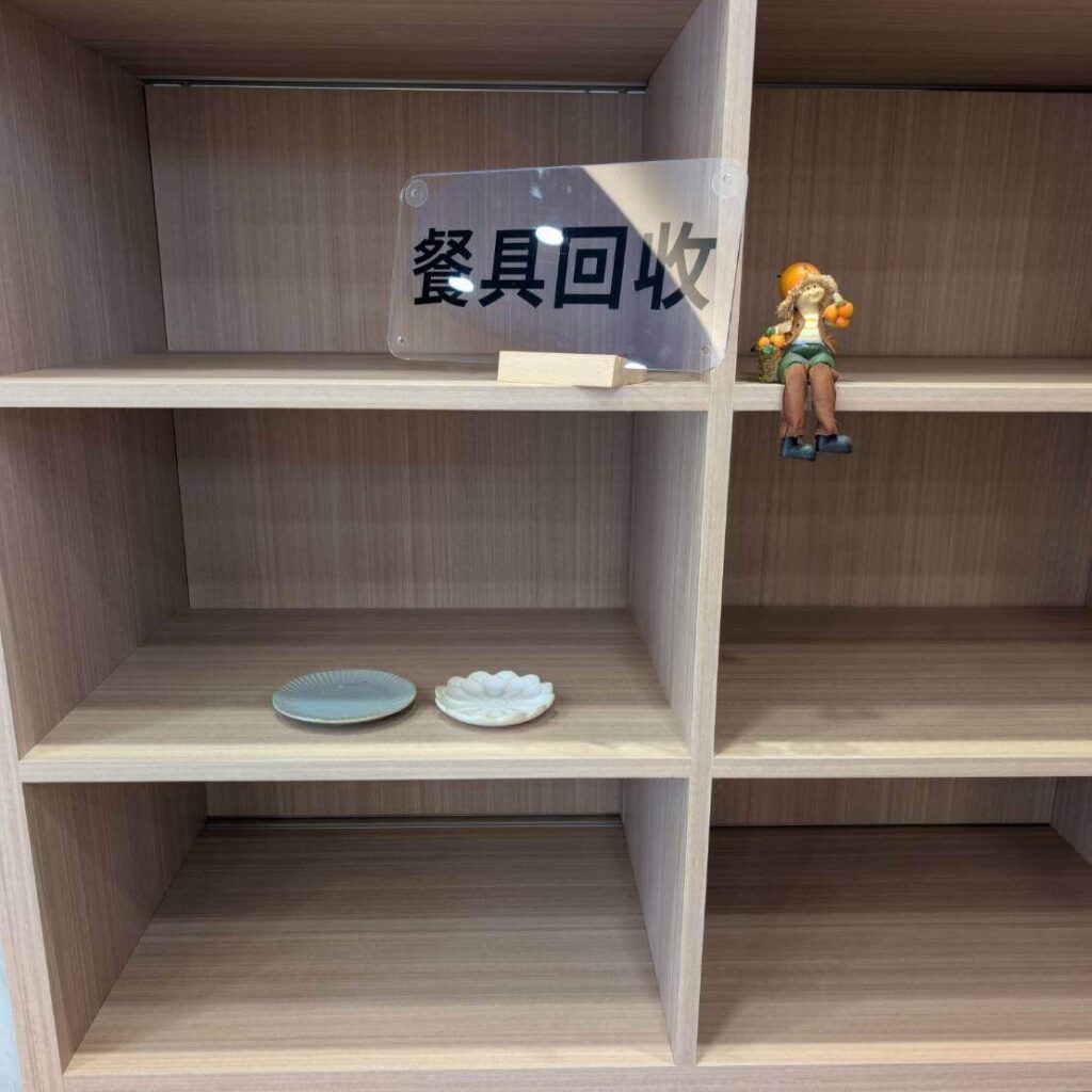 【瑞穗美食】「縱谷耘穗咖啡廳」的餐具回收區，木質櫃體上放有空盤與「餐具回收」標示牌，旁邊擺著一個可愛的農夫造型小擺飾，整體乾淨整齊。