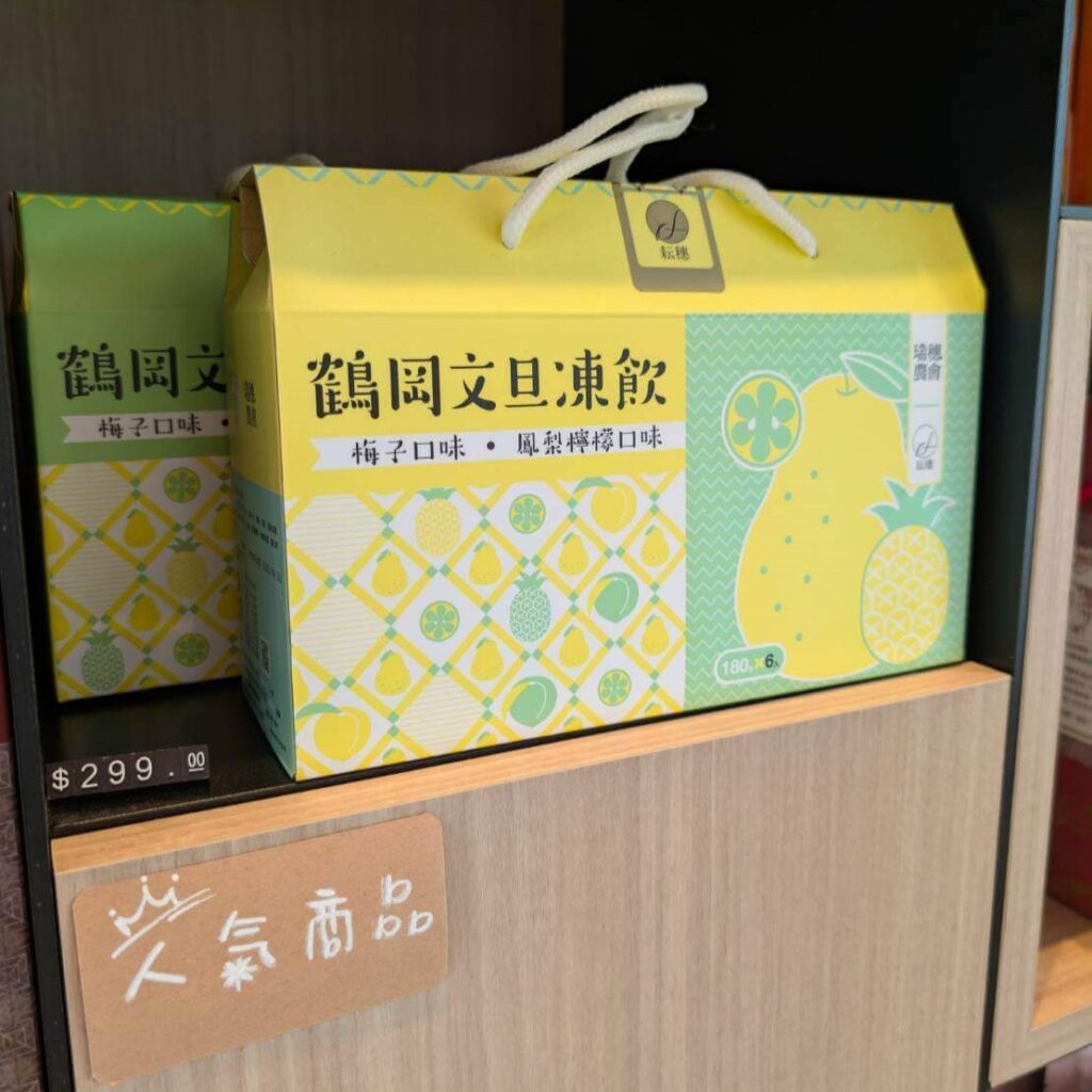 【瑞穗美食】「縱谷耘穗咖啡廳」販售的熱門商品「鶴岡文旦凍飲」，包裝以黃色與綠色設計，主打梅子與鳳梨檸檬口味，標示價格為299元，陳列於木質架上並附有手寫小牌「人氣商品」。