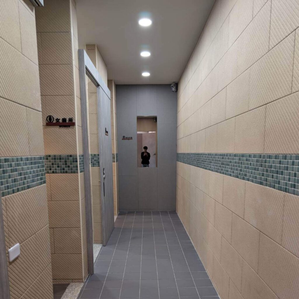 【瑞穗美食】「縱谷耘穗咖啡廳」的廁所走廊，牆面採用米色與藍綠色拼接磁磚設計，燈光明亮，空間整潔，盡頭設有鏡面與標示清楚的男女廁門。