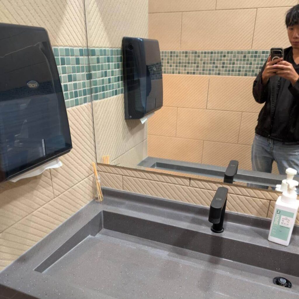 【瑞穗美食】「縱谷耘穗咖啡廳」的洗手間一角，設有灰色石質洗手台與黑色龍頭，牆面為米色與綠色馬賽克磁磚設計，整體乾淨明亮。鏡中可見生活口袋創作者拍攝的倒影，檯面上放有洗手乳與竹籤罐，展現整潔細緻的空間氛圍。