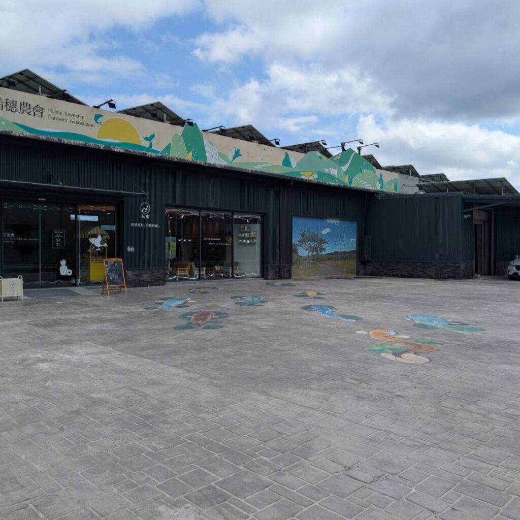 【瑞穗美食】「縱谷耘穗咖啡廳」與「琍穗農會」建築外景全貌，前方為寬闊的停車空間，地面有繽紛魚群彩繪，屋頂設有太陽能板，整體融合自然與在地農村風情。