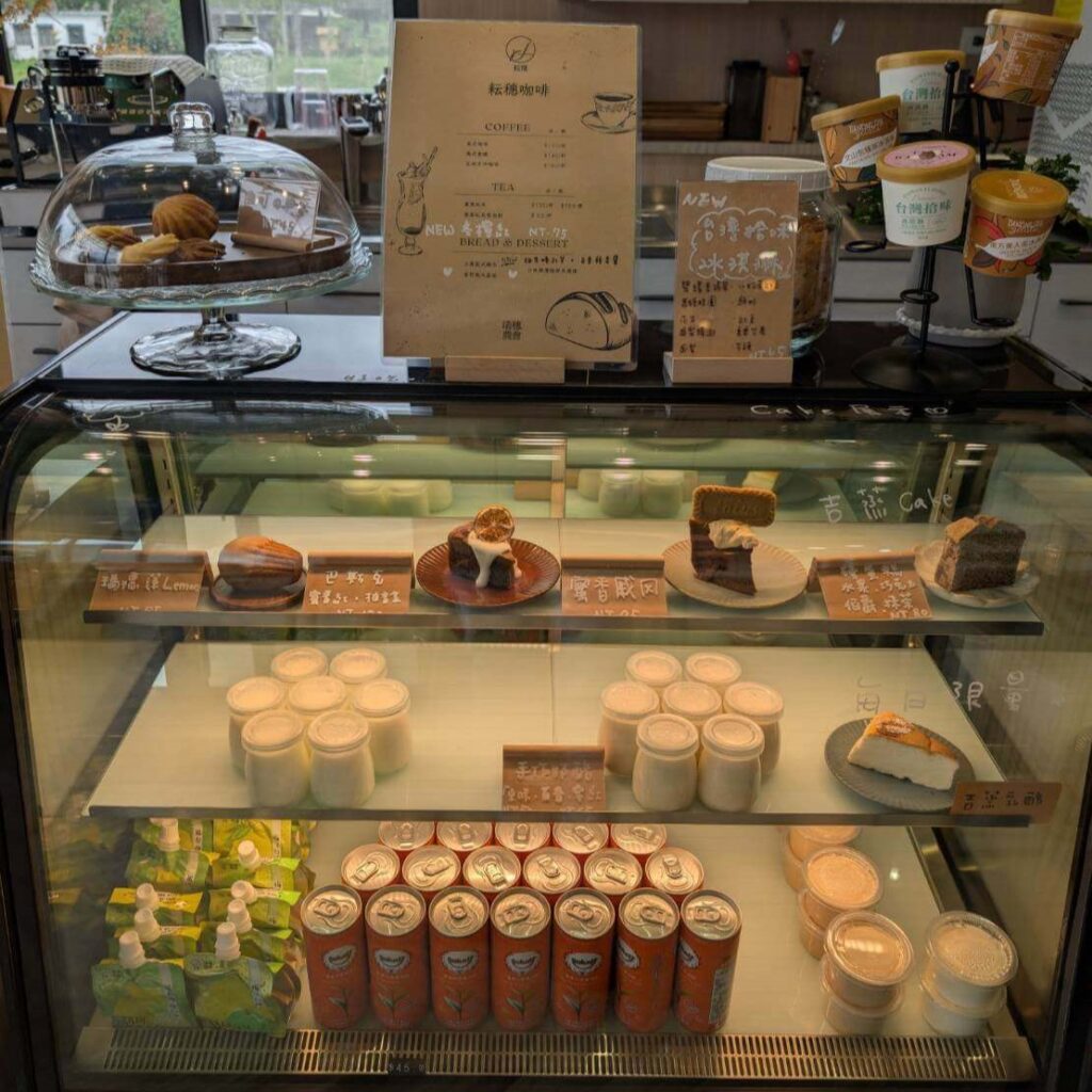 【瑞穗美食】"縱谷耘穗咖啡廳精選示意圖"「縱谷耘穗咖啡廳」的甜點展示櫃，內有各式蛋糕與布丁，包含檸檬蛋糕、巴斯克乳酪、提拉米蘇、戚風蛋糕與每日限量甜點，上層放有咖啡菜單與冰淇淋杯。整體氛圍乾淨明亮，櫃台後方可見咖啡機與餐具。