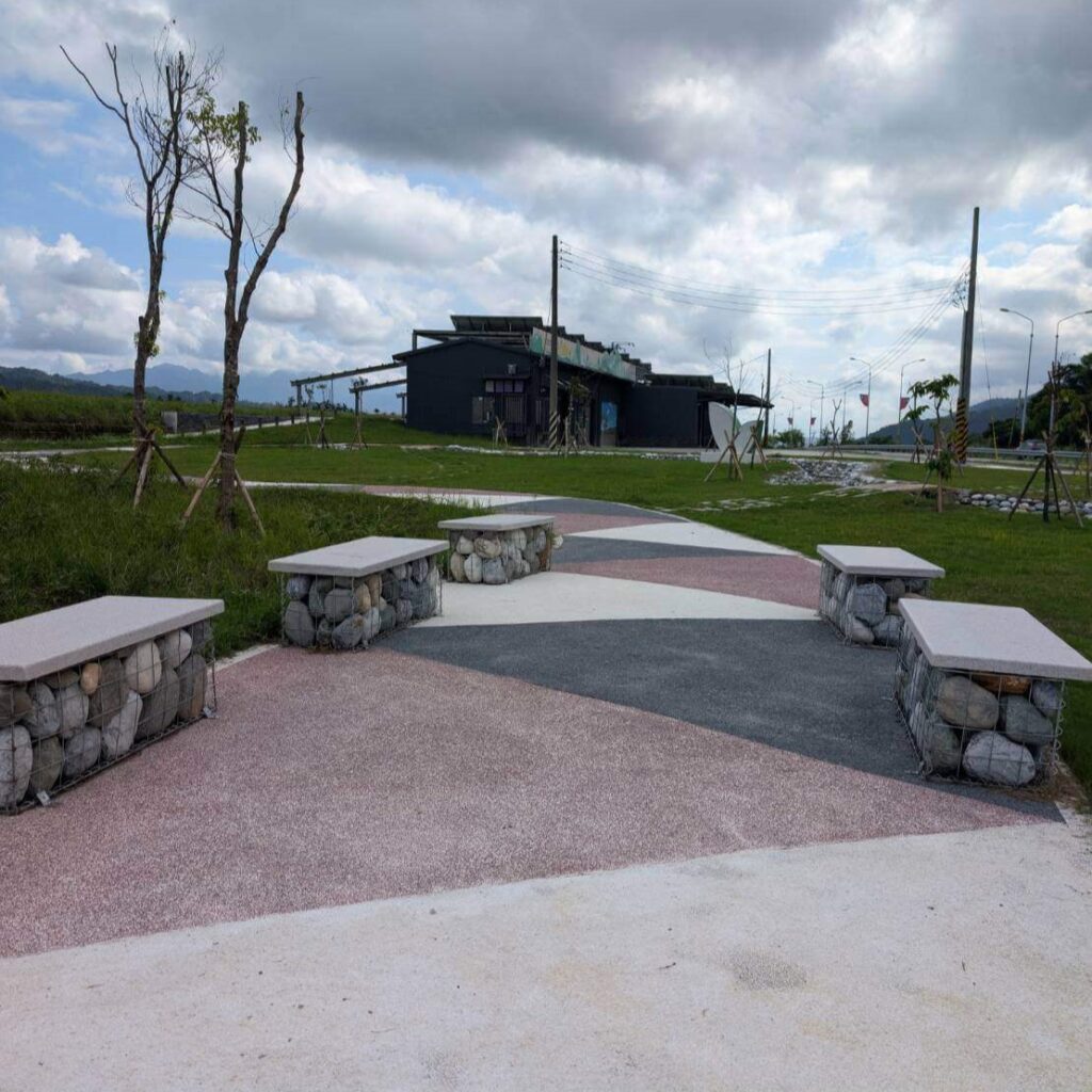 【瑞穗美食】「縱谷耘穗咖啡廳」外部景觀步道，設有石砌長凳與彩色鋪面通道，連接咖啡廳主建築，背景可見藍天白雲與綠意山景，環境開闊且充滿自然氣息。