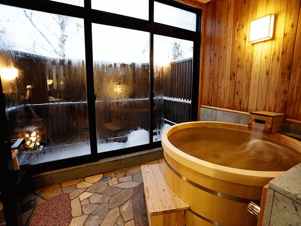 【網走住宿】「能取之莊 Kagari 屋」的客房內私人溫泉浴池，以木桶浴槽與石材地板打造，窗外可見冬日雪景，充滿溫泉旅館的靜謐氛圍。