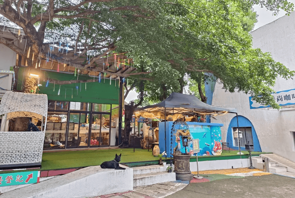 【六龜住宿】高雄六龜【道時尚悠然山莊】園區入口與尚咖啡館外觀，樹蔭下的戶外座位與繽紛裝飾營造度假氛圍，一旁黑狗悠閒休憩，展現鄉村自然風情。