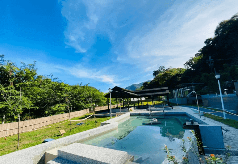 【六龜住宿】高雄六龜【遠山望月溫泉風呂露營區】露天溫泉池白天景色，湯池環境寬敞，周圍綠樹成蔭，遠方山景映照水面，氛圍悠閒靜謐。