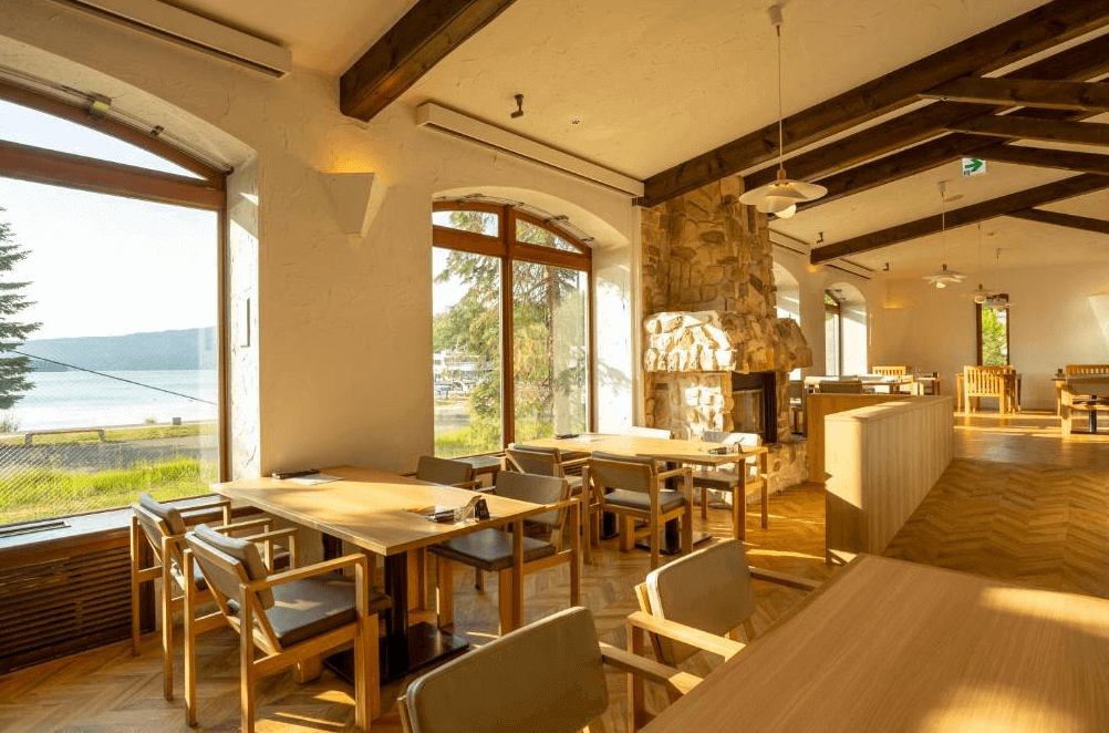 【釧路住宿】阿寒之森鶴雅度假村湖畔餐廳，陽光灑落於木質桌椅與石造壁爐之間，窗外可見阿寒湖景色，營造溫馨自然的用餐環境。