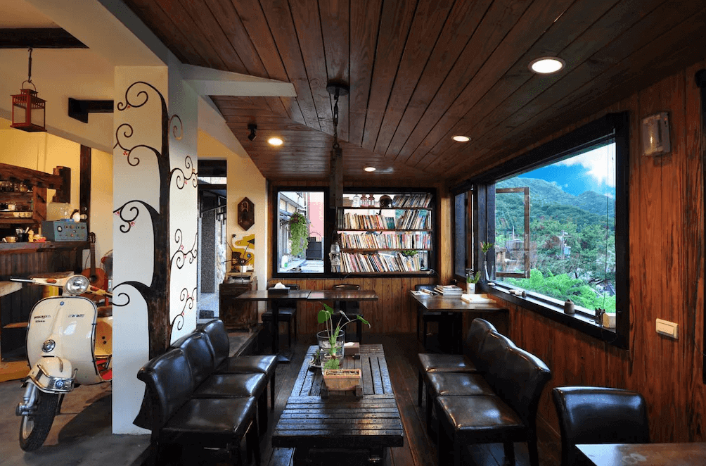 【瑞芳住宿】陽光味宿｜公共交誼廳結合書香與咖啡氛圍，木質空間內設有書櫃與大窗景，可一邊閱讀一邊欣賞九份山城景緻。