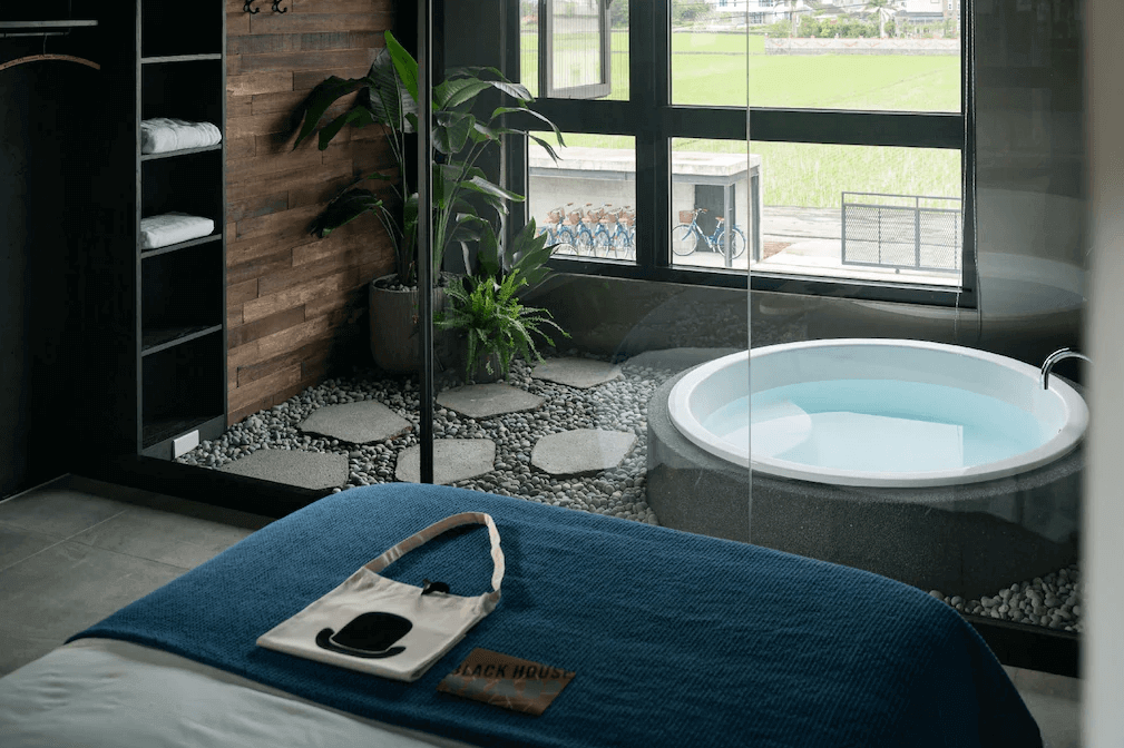【頭城住宿】宜蘭黑宅 Black House 房內景觀浴池設計，圓形石造浴缸臨窗而設，可欣賞稻田景色，室內融合木質牆面與綠植造景。