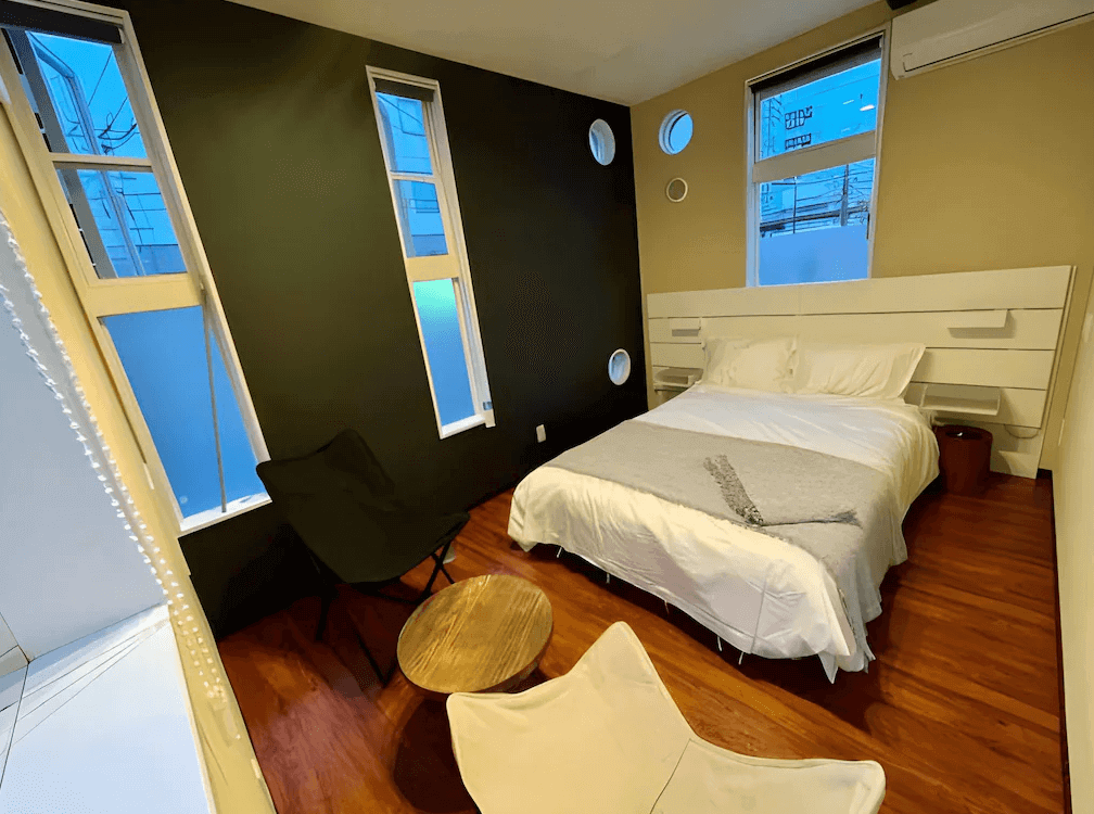 【奄美大島住宿】「En-Hostel & Café Bar 雙人客房：設計感強烈的臥室空間，大片縱向窗戶搭配深色牆面與木質地板，配置一張雙人床與舒適休憩座椅。」