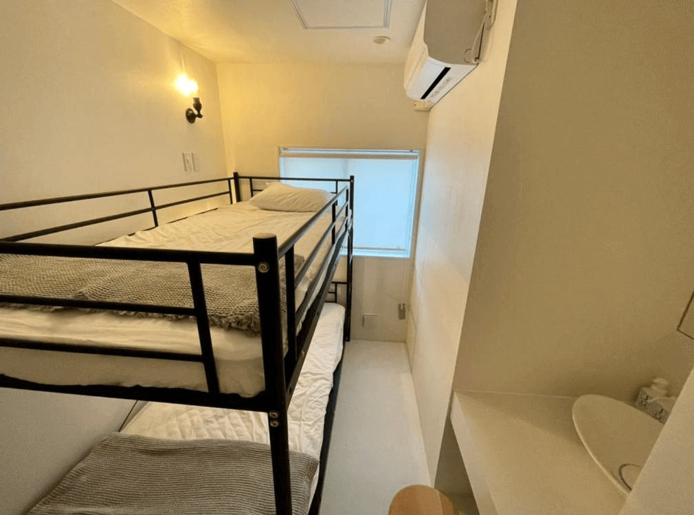 【奄美大島住宿】「En-Hostel & Café Bar 四人上下舖房型：白色系簡約空間中設有金屬上下舖床架，旁邊為小型洗手台，整潔明亮。」