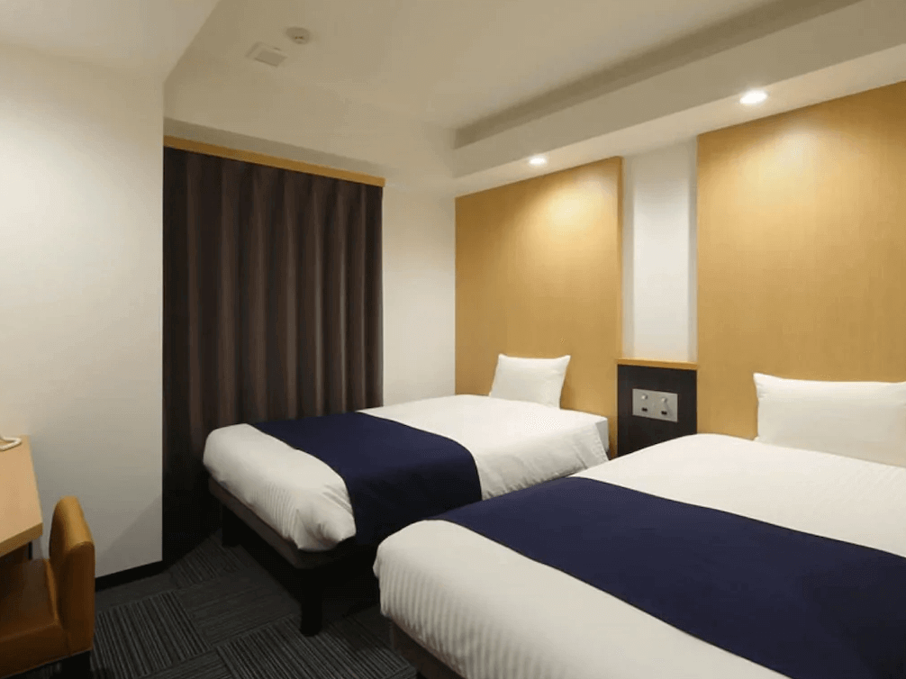 【奄美大島住宿】「Hotel West Court Amami 2 雙床房：簡約溫暖的木質調客房設有兩張單人床，搭配深藍色床尾巾與柔和燈光，空間舒適整潔。」