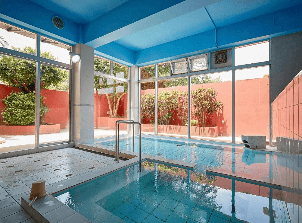 【讀谷村住宿】「Murasakimura 飯店的室內溫泉池，明亮採光搭配藍色池水與大片落地窗，可欣賞庭院綠意，提供旅客放鬆療癒的泡湯環境。」