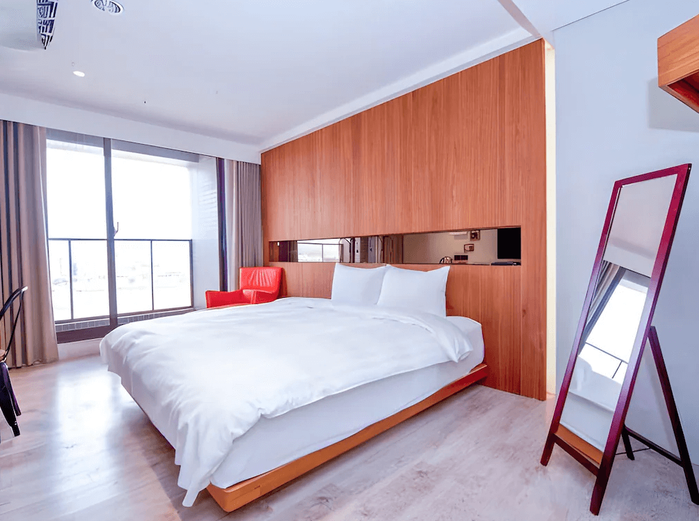 【台中火車站住宿】「old school 行旅・田中央旅店的雙人房，採用溫潤木質設計與大片落地窗，搭配純白床鋪與紅色休閒椅，整體風格明亮簡約。」