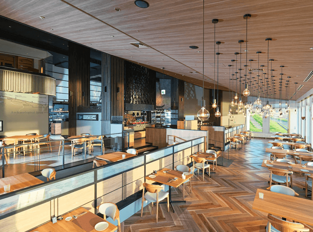 【北見住宿】佐呂間湖鶴雅度假飯店的餐廳環境，挑高木質天花搭配吊燈設計，大片玻璃窗可眺望戶外綠景，整體氣氛溫馨又具現代感。