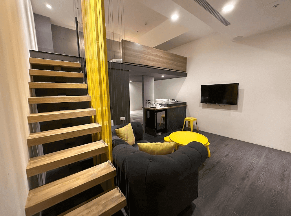 【台中火車站住宿】「凝萃文旅台中車站店的樓中樓房型，室內以黃灰配色呈現，設有階梯通往上層休憩空間，並配置現代風格沙發、茶几及電視，營造寬敞舒適的住宿氛圍。」