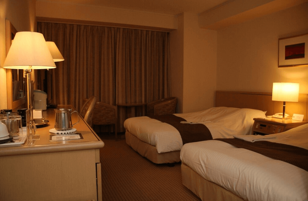 【北見住宿】北見皮爾森酒店客房：溫馨雙床房設計，柔和燈光搭配米色調裝潢，設有書桌、熱水壺與茶具設備，窗邊擺放兩張休閒椅與小圓桌，營造舒適住宿氛圍。