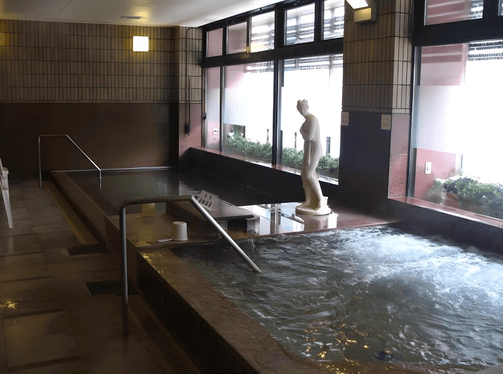 【北見住宿】北見皮爾森酒店溫泉浴池：館內公共溫泉浴池區，採用石質設計，窗邊設有雕像裝飾，空間寬敞明亮，提供溫泉泡湯與按摩水療設施。