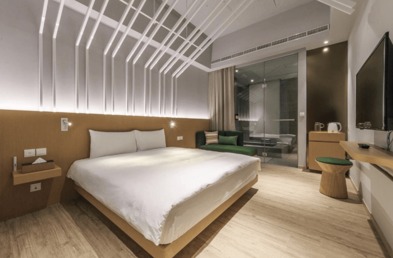 【宜蘭溫泉飯店】「品文旅現代簡約雙人房，房內以溫暖木質與白色燈條裝飾天花板為設計亮點，配置大床、綠色沙發與玻璃隔間浴室，整體明亮舒適。」
