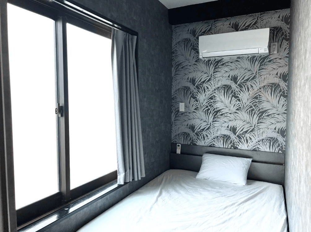 【奄美大島住宿】「奄美 JOOOY Hotel 的單人房，房內以灰色調與葉紋壁紙打造現代風格，配有大窗與獨立冷氣的舒適睡眠空間。」