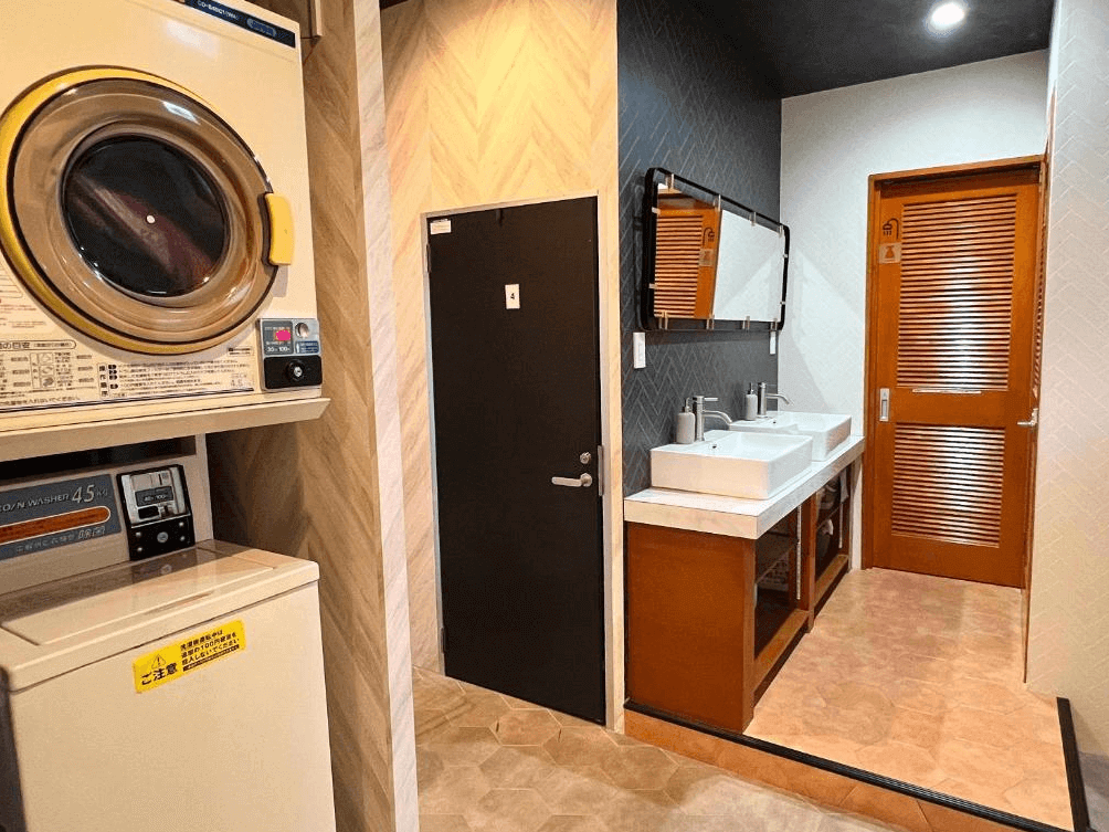 【奄美大島住宿】「奄美 JOOOY Hotel 的共用洗手區與洗衣設備，包含洗手台、鏡面、烘衣機與洗衣機，提供長住旅客便利的生活機能。」