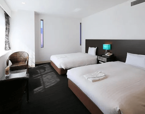 【奄美大島住宿】「奄美里貝斯特酒店（Libest Inn Amami）雙床房角度展示：房內以沉穩色調搭配藍色檯燈與籐椅，整體呈現簡約舒適的住宿氛圍。」