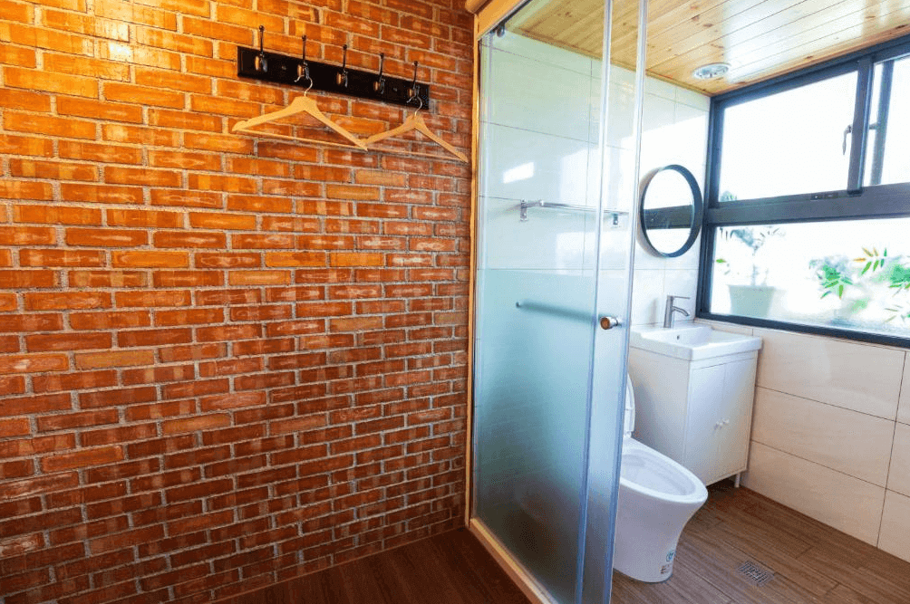 【大溪住宿】小巷內客棧 Hostel Alley Inn 浴室空間，紅磚牆面搭配木質天花板與大面採光窗，乾濕分離設計並設有圓形鏡面與白色洗手台，風格溫馨自然。