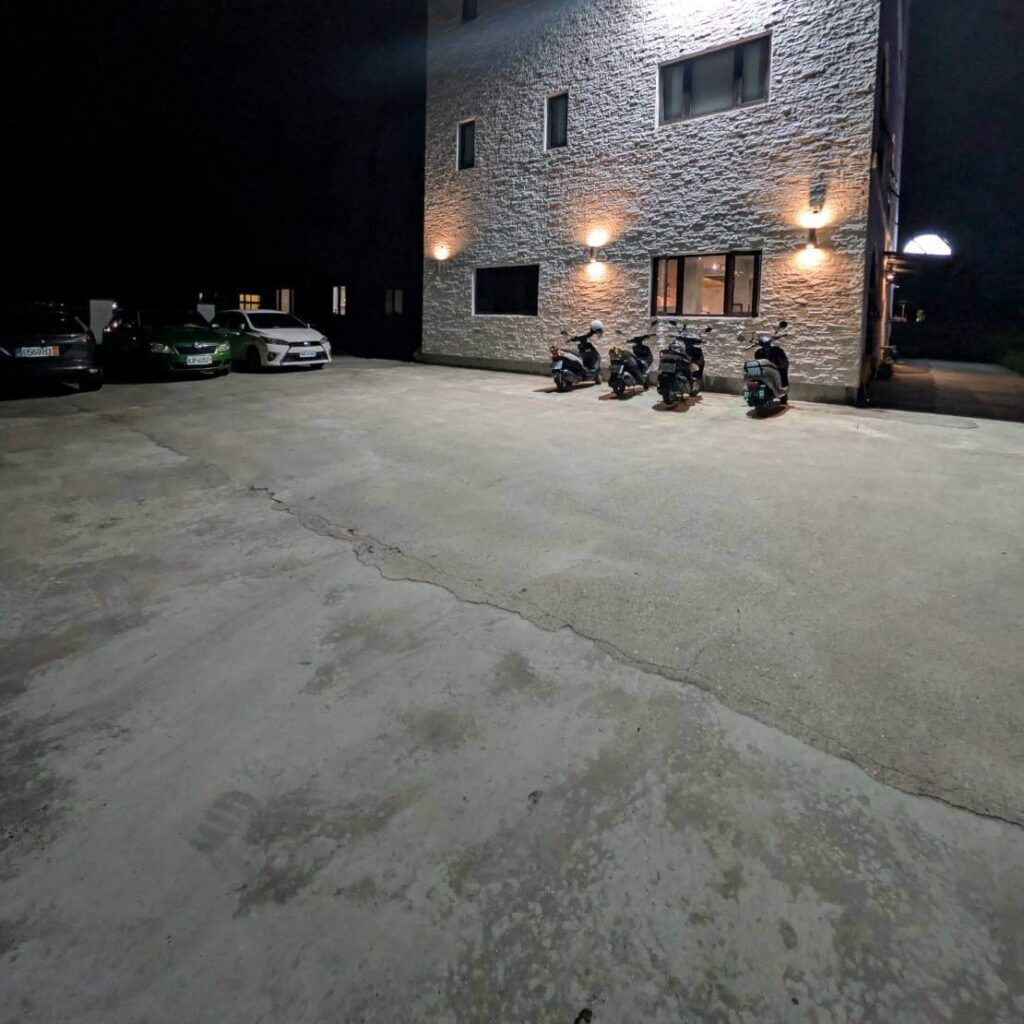 【山杉青旅】「山杉青旅戶外停車區夜景，寬敞的水泥空地上停放多台汽車與機車，旅館外牆以白色石紋設計搭配暖色壁燈照明，展現旅宿提供方便安全的停車環境。」