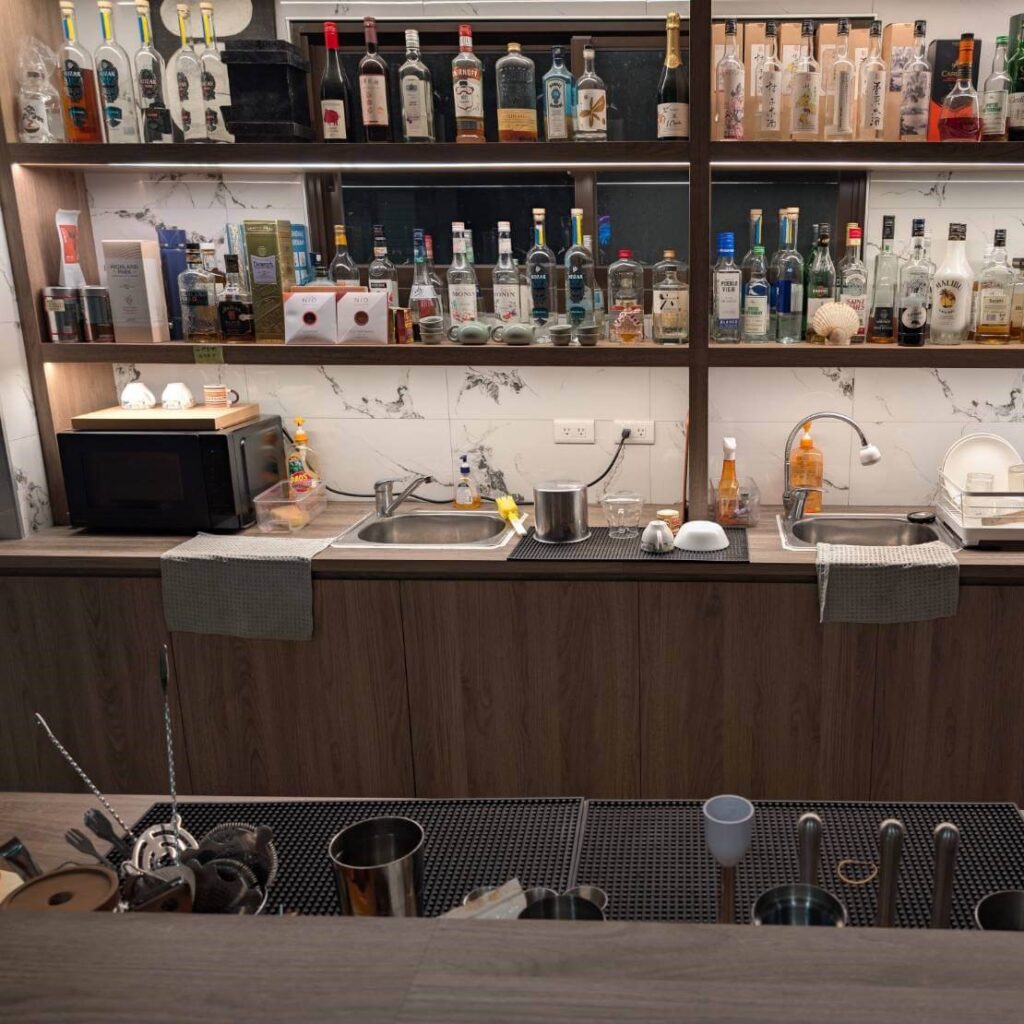 【山杉青旅】「山杉青旅吧檯的後場工作區，牆面層架整齊擺放大量酒瓶與飲品用品，下方設有微波爐、水槽與咖啡用具，檯面上散放杯具與調飲工具，呈現旅宿提供飲品服務的備作空間。」