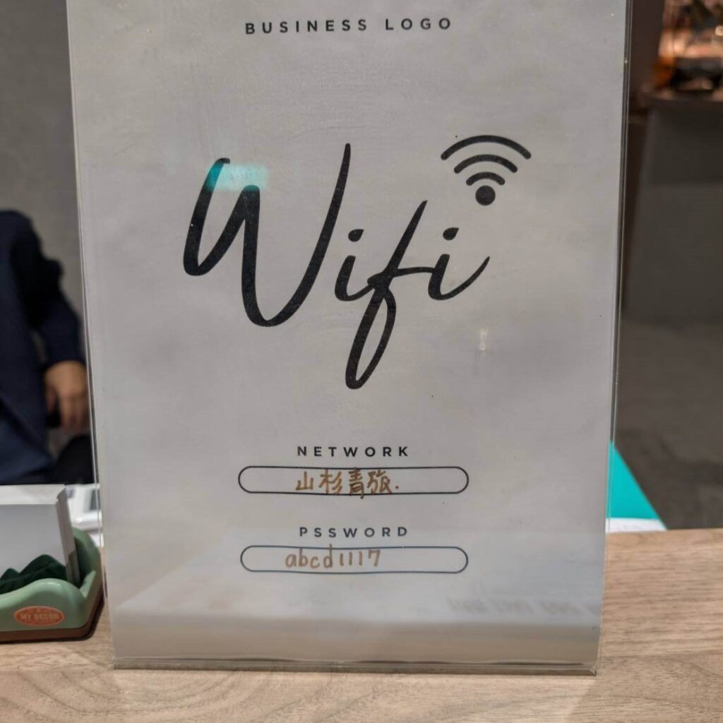 【山杉青旅】「山杉青旅櫃台上放置的 Wi-Fi 資訊立牌，牌面上以大字寫著 Wifi，並標示網路名稱『山杉青旅』與密碼『abcd1117』，背景為室內接待空間。」