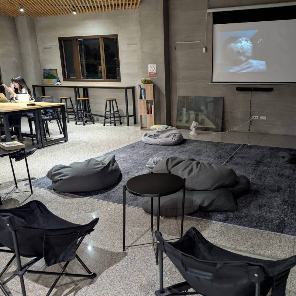 【山杉青旅】"山杉青旅住宿精選示意圖"「山杉青旅大廳的公共空間，地上鋪有大面積深色地毯，擺放多個灰色懶骨頭與摺疊椅，前方投影幕正播放影片，右側牆面有投影設備，左側為工作桌與高腳椅，是旅客可休息、觀影與交流的舒適共享區域。」