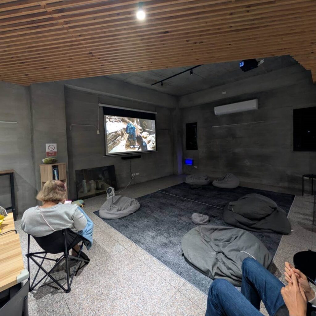 【山杉青旅】「山杉青旅大廳的影視放映區，灰色地毯上散放多個懶骨頭座墊，旅客正坐在折疊椅上觀看投影幕播放的影片，周圍為水泥質感牆面與木質天花板，營造舒適放鬆的觀影空間。」