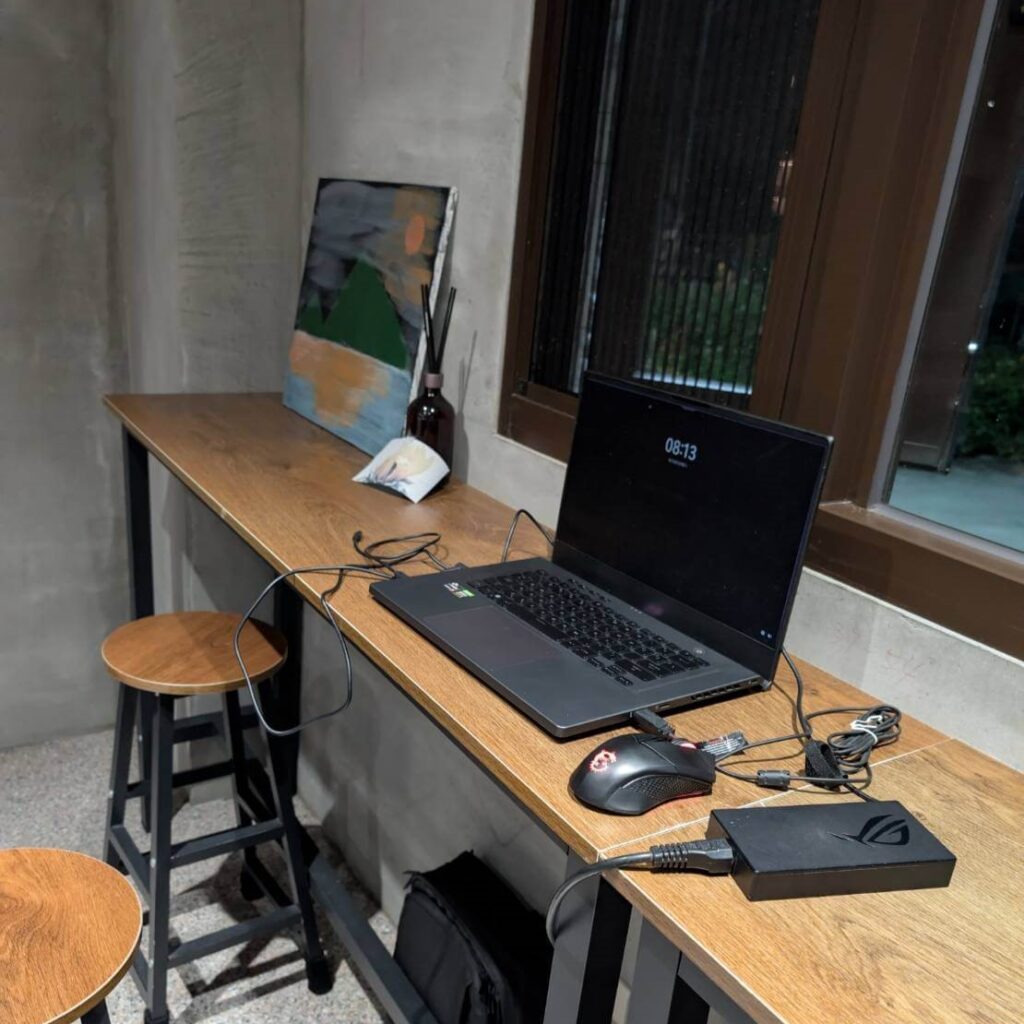 【山杉青旅】「山杉青旅窗邊的高腳座位區，一台筆電與滑鼠放在木質長桌上，旁邊擺放抽象風格畫作與擴香瓶，搭配金屬腳架高腳椅，呈現適合旅客工作與放鬆的文青風公共空間。」