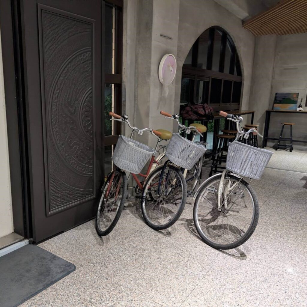 【山杉青旅】「山杉青旅大廳入口旁擺放三台供房客使用的腳踏車，車籃與車身設計簡約，背景為室內水泥風格牆面與木質高腳桌椅，展現旅宿貼心提供代步工具的休閒氛圍。」