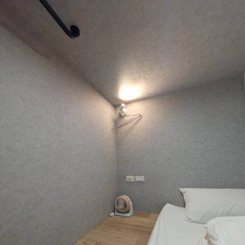 【山杉青旅】「山杉青旅膠囊房內部空間，灰色隔音材質牆面搭配溫暖的床頭燈，牆上設有金屬衣架與插座，下方放置一台小型循環扇，整體呈現簡潔安靜的睡眠環境。」