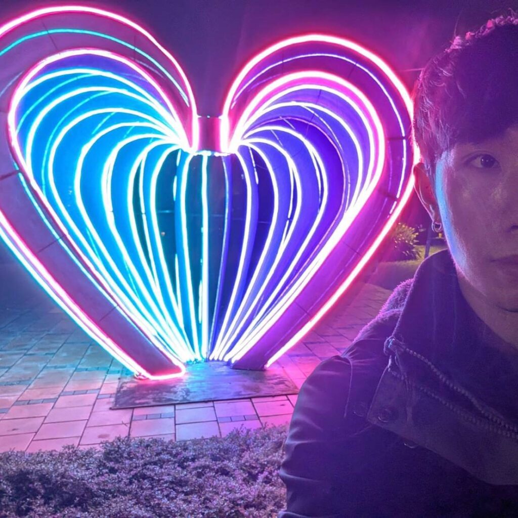 【山杉青旅】「山杉青旅門口對面的三民濕地公園夜景，一位旅客在彩色霓虹燈打造的巨大愛心裝置前自拍，燈光以藍、紫、粉色漸層呈現，形成浪漫繽紛的打卡景點氛圍。」