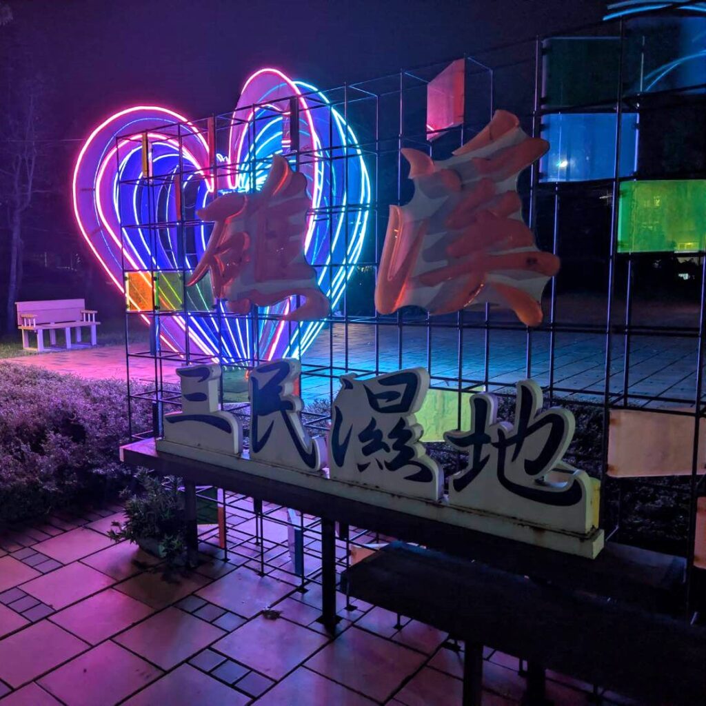 【山杉青旅】「山杉青旅門口對面的三民濕地公園夜景，園區入口矗立著寫有『三民濕地』與『雄戀』的大型裝置字樣，後方以彩色霓虹心形燈飾點亮夜空，營造繽紛浪漫的散步氛圍。」