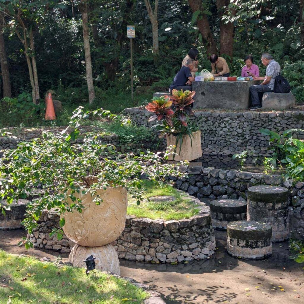 【山杉青旅】「山杉青旅周邊景點森林風呂旁的休憩區，多位旅客坐在石造桌椅上聚會用餐；前景為層層堆疊的石牆造景、植栽盆景與淺水步道，呈現自然與人文融合的悠閒氛圍。」