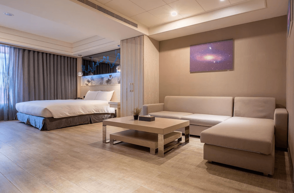【台中火車站住宿】「承攜行旅台中自由館的寬敞客房，一旁設有大面 L 型沙發與茶几，柔和燈光營造溫馨氛圍，房內以木質調與太空主題掛畫呈現雅緻設計。」