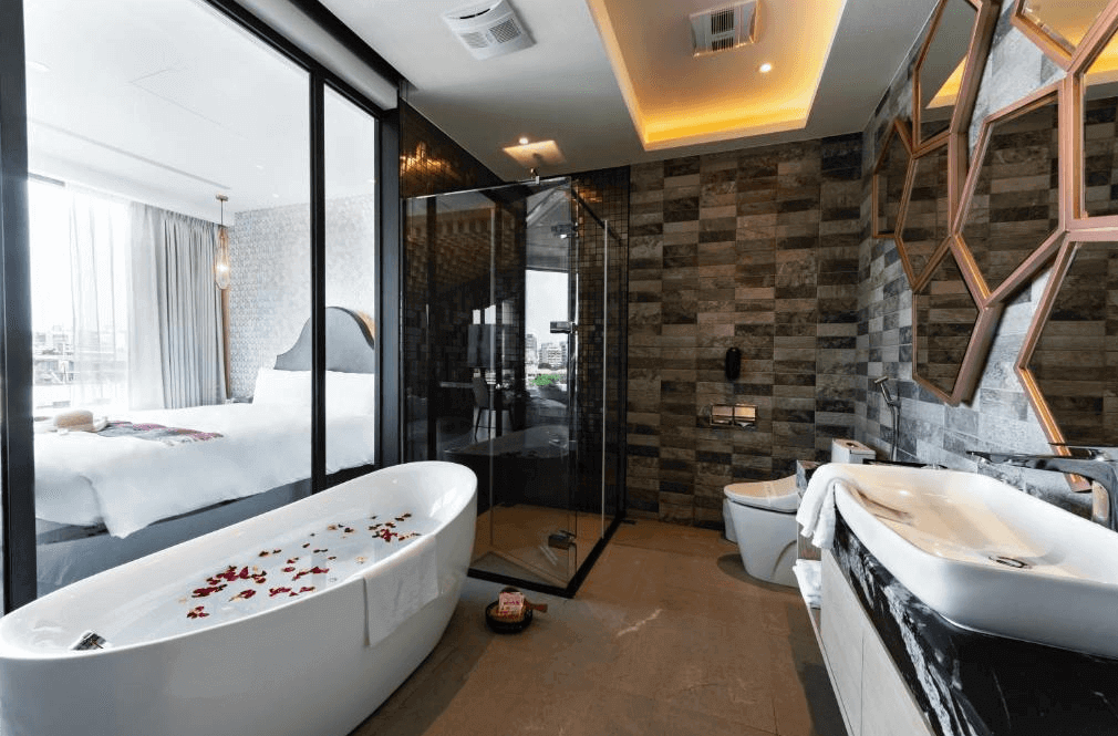【台中火車站住宿】「挪威森林台中文創館的豪華浴室，配有獨立浴缸、淋浴間與馬桶區，搭配深色石紋牆面與六角鏡面裝飾，可從透明隔間看見臥室與窗外景觀，整體設計質感十足。」
