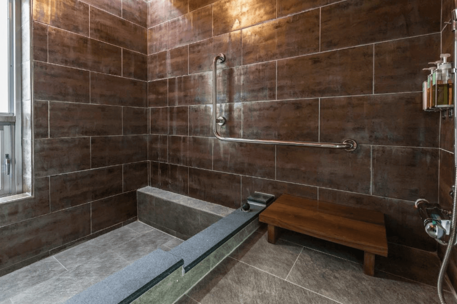 【宜蘭溫泉飯店】「有朋會館浴室空間，採用深色磚牆與大型溫泉浴池設計，設有安全扶手與木質置物椅，整體風格沉穩質樸，具溫暖療癒感。」