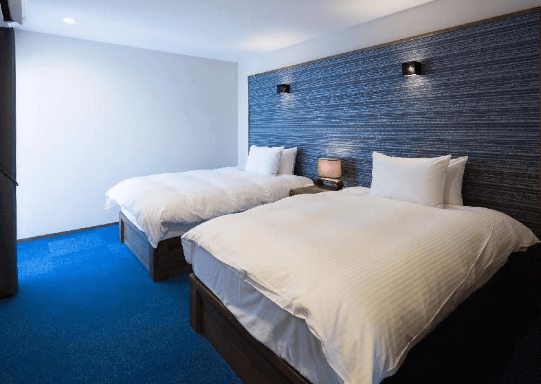 【讀谷村住宿】「沖繩本島T客房公寓的雙床房型，房內配置兩張柔軟的大床，搭配簡約白色寢具與深色紋理牆面，地毯採亮藍色設計，整體呈現溫馨又清爽的住宿空間。」