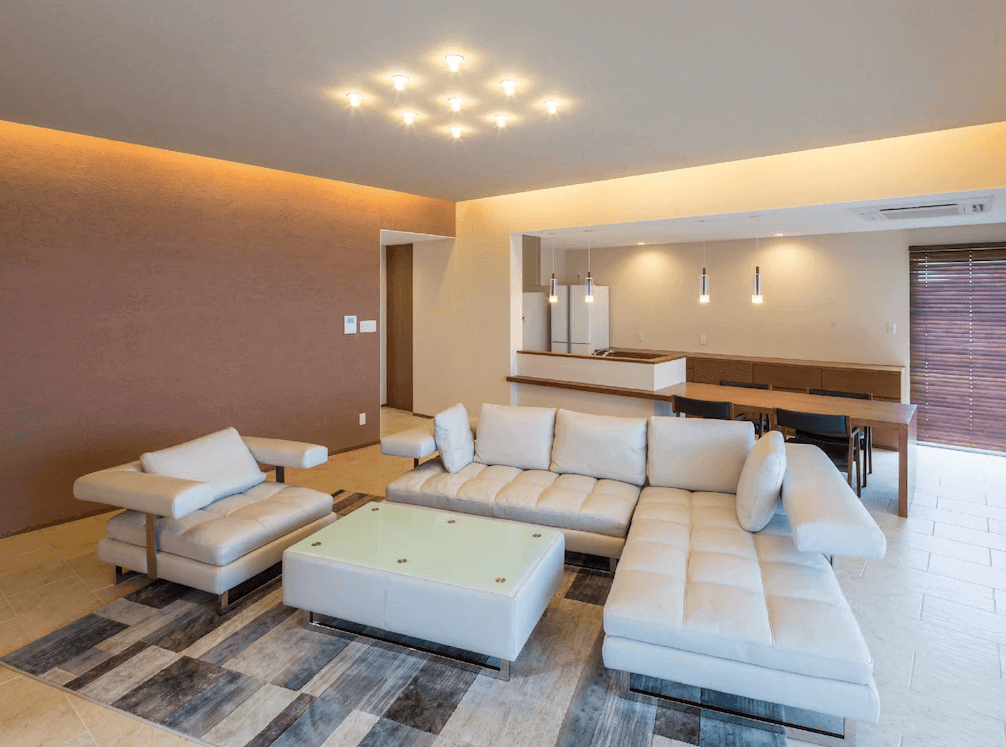 【讀谷村住宿】「沖繩本島T客房公寓寬敞明亮的客廳空間，配有大型米色L型沙發、玻璃茶几與溫暖燈光，後方連結開放式餐廳與廚房，營造舒適的度假居家氛圍。」