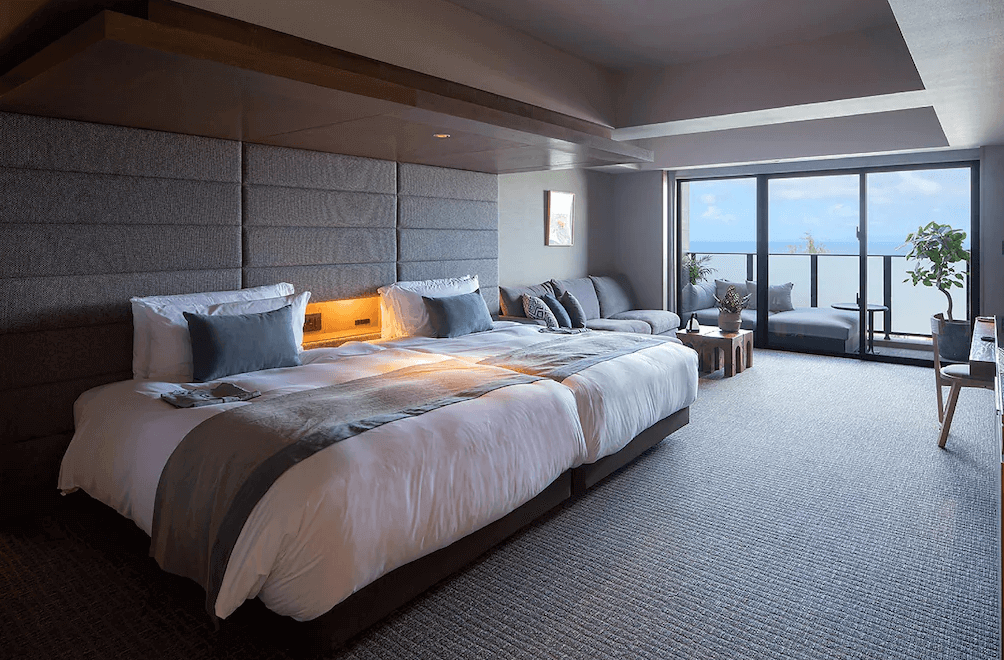 【讀谷村住宿】「沖繩讀谷 GLAMDAY STYLE 酒店及度假村的海景豪華客房，寬敞大床搭配灰色系設計，落地窗外可見藍色海景，整體呈現靜謐且高雅的度假氛圍。」