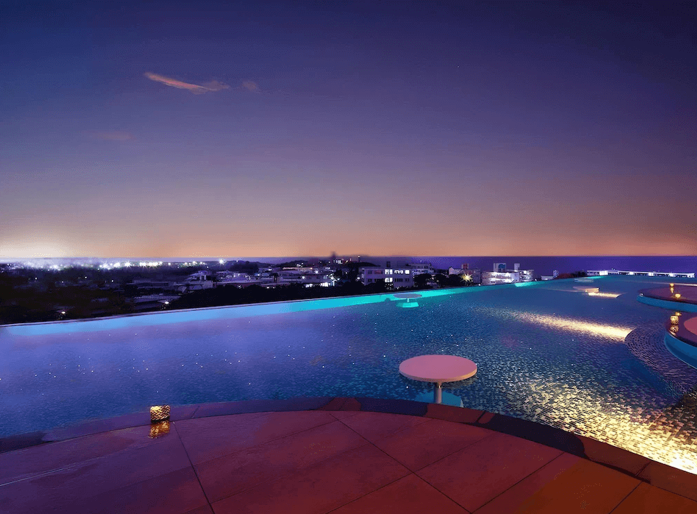 【讀谷村住宿】「沖繩讀谷 GLAMDAY STYLE 酒店及度假村的無邊際泳池夜景，池水倒映城市燈光與海平面，營造浪漫且奢華的度假夜晚。」